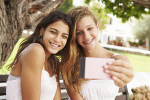 Two Teenage Girls Sitting On Bench Taking Selfie In Park
